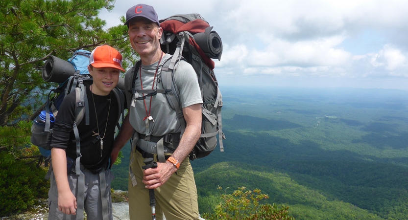 A parent and child smile for a photo on an overlook high above a green mountainous area. Both are wearing backpacks.
